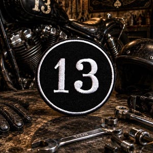 Op de afbeelding: Een ronde, zwart geborduurde patch met een groot wit cijfer 13 in het midden, omringd door een dunne witte rand. De patch wordt getoond tegen een donkere, industrieel ogende achtergrond met motoronderdelen en metalen gereedschappen.