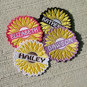 May include: Embroidered sunflower patches in yellow, pink, black, and white. Each patch features a name: Elizabeth, Kather, Dawson, and Hailey, embroidered on a white or colored banner across the center. The patches are arranged on a textured surface.