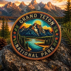May include: Embroidered patch featuring a scenic landscape of the Grand Teton National Park. The circular patch displays mountains, a lake, and the text "GRAND TETON NATIONAL PARK" in a decorative font. The patch is bordered by a floral design.