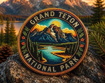 May include: Circular embroidered patch depicting the Grand Teton National Park. The design includes mountains, a lake, and the text "GRAND TETON NATIONAL PARK". The patch is framed by a floral border. The colours are vibrant.