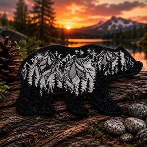 May include: A black embroidered patch shaped like a bear, featuring a white stitched design of a mountain range, pine trees, and a crescent moon with stars. The patch is displayed on a textured wooden surface with a pinecone and small stones.