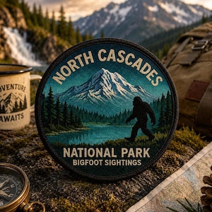 May include: Circular patch with the text "North Cascades National Park Bigfoot Sightings" featuring a mountain scene and a silhouette of Bigfoot. The patch is surrounded by a forest, a lake, and a compass.