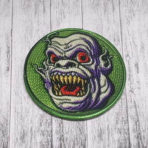 Könnte beinhalten: Ein runder, grüner gestickter Aufnäher mit einem weißen und violetten Monstergesicht mit roten Augen und scharfen Zähnen. Das Monster knurrt mit offenem Maul.