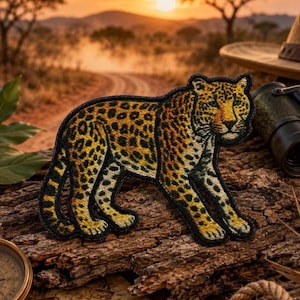 May include: An embroidered patch shaped like a leopard in profile, featuring a golden-yellow coat with black spots, a white underbelly, and a dark stitched border. The leopard is depicted in a standing position with its head turned towards the viewer.