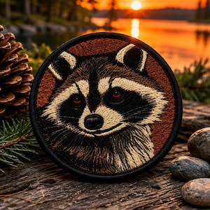 May include: A circular embroidered patch featuring a detailed raccoon face with black, white, and brown thread. The raccoon has distinct dark eye patches, a black nose, and red eyes. The patch has a solid black stitched border edge.