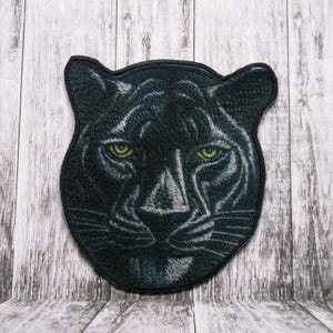 Peut inclure: Patch brodé représentant la tête d'une panthère noire avec des yeux verts. Le patch est en tissu et a un contour noir.