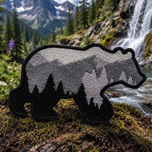 Könnte beinhalten: Ein gestickter Aufnäher in Form eines laufenden Bären. Das Design zeigt eine schwarze Silhouette des unteren Körperbereichs und der Beine, die in eine grau-weiße Landschaft aus Tannenbäumen und Berggipfeln im oberen Körperbereich übergeht.