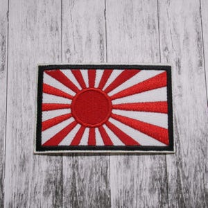 May include: A red and white embroidered patch with a red circle in the center and 16 white rays radiating out from the circle. The patch is surrounded by a black border and a white border.