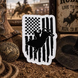 May include: A rectangular embroidered patch featuring a black and white design of a bull rider silhouette superimposed over a stylized American flag with stars and stripes, finished with a scalloped white border edge.