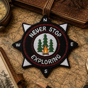 May include: Black and white embroidered patch in a compass rose design with the text "NEVER STOP EXPLORING". The patch features a central image of green trees and a campfire. The patch is surrounded by a red border.