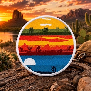 Può includere: Una toppa ricamata circolare raffigurante un paesaggio desertico con cielo giallo, cactus verdi, montagne rosse e un primo piano blu con una luna bianca. Il design è bordato da un bordo cucito bianco, che mostra una vivace scena al tramonto.