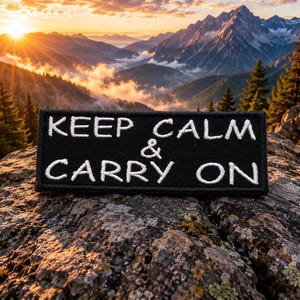 May include: A rectangular black embroidered patch featuring the white stitched text KEEP CALM & CARRY ON. The patch has a clean, straight-edged border and is positioned horizontally, displaying clear, legible lettering against the dark fabric background.