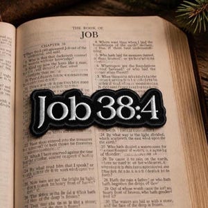 May include: A black and white embroidered patch with the text "Job 38:4" is displayed on an open book. The patch has a black border and white lettering. The book is open to a chapter from the Book of Job.