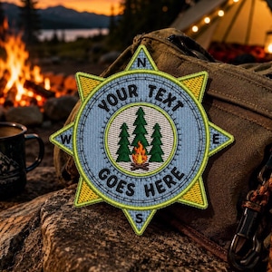 May include: Embroidered patch with a compass design. The patch features a light blue background with the text "YOUR TEXT GOES HERE" and a central image of pine trees and a campfire. The compass points are marked with N, E, S, and W. The patch is yellow and green.