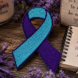 May include: A teal and purple ribbon with embroidered details, resting on a wooden surface. A small notebook with the words "hope, healing, strength, love" is next to the ribbon. A candle and a sign with text are also in the image.