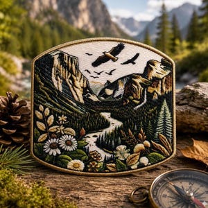 May include: Embroidered patch featuring a mountain landscape with a river, trees, and flying birds. The patch has a cream-colored background with a gold border. The design includes detailed stitching of flowers and foliage.