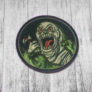Könnte beinhalten: Runder, gestickter Aufnäher mit einem grünen und weißen Monster mit roten Augen und offenem Mund. Das Monster ist von Grün umgeben und vor einem dunkelgrünen Hintergrund mit einem lila Rand platziert. Für Kleidung.