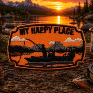 Puede incluir: Un parche bordado naranja con borde negro que presenta la silueta de una persona pescando desde un bote en un lago con montañas al fondo. El texto MY HAPPY PLACE está bordado en negro en la parte superior del parche rectangular.