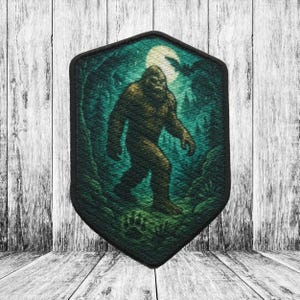May include: A Bigfoot patch depicting a forest scene. The patch shows a brown Bigfoot walking through a dark green forest under a full moon. The patch has a black border and a unique shape.