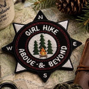 Op de afbeelding: Zwart en wit geborduurde patch met de tekst "GIRL HIKE ABOVE & BEYOND" in een kompasontwerp. De patch heeft een kampvuur en bomen. De patch ligt op een kaart met een kompas en een mok.