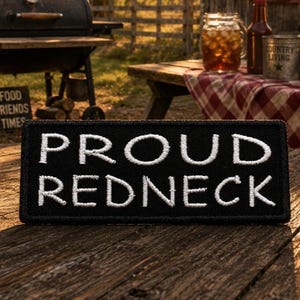 May include: A rectangular black embroidered patch featuring the white text PROUD REDNECK in a bold, sans-serif font. The patch has a stitched border and is positioned horizontally on a weathered wooden surface with a visible texture.