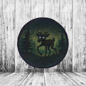 May include: A round patch featuring a deer silhouette with large eyes, set against a forest backdrop. The deer is dark brown, and the forest is a mix of green and dark green. The patch has a black border and is set against a white wooden background.