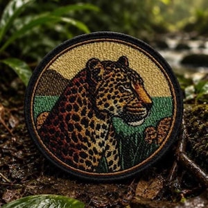 May include: A circular embroidered patch featuring a detailed leopard profile in shades of gold, brown, and black. The leopard is set against a background of a light tan sky and a green landscape, all enclosed within a thick black stitched border.