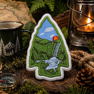 Peut inclure: Écusson brodé en forme de flèche, représentant un paysage de montagne avec une cascade, une rivière et un coucher de soleil. L'écusson a une bordure blanche et est posé sur un fond d'équipement de camping, dont une lanterne, une boussole et une tasse.