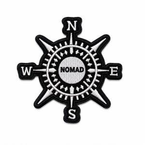 May include: A black and white embroidered patch featuring a compass rose design. The center circle contains the text NOMAD in bold letters. Four cardinal directions N, S, E, and W are positioned at the tips of the compass points in white.