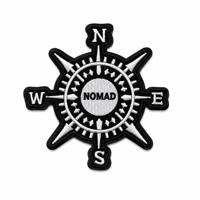 May include: A black and white embroidered patch featuring a compass rose design. The center circle contains the text NOMAD in bold letters. Four cardinal directions N, S, E, and W are positioned at the tips of the compass points in white.