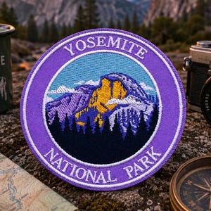 May include: A round, embroidered patch with the text "YOSEMITE NATIONAL PARK" in white on a purple border. The patch features a mountain landscape in blue, yellow, and purple. A green enamel mug and a vintage camera are in the background.