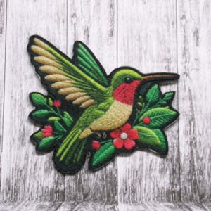 May include: Green and yellow hummingbird with red accents, perched on a green leafy branch with red flowers. The hummingbird is in flight with its wings spread out.
