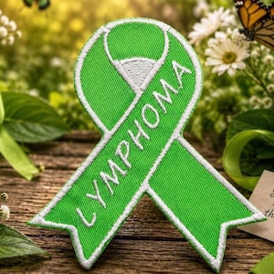 May include: A green ribbon with the word "LYMPHOMA" embroidered in white. The ribbon is surrounded by flowers and greenery, with a butterfly in the background. A small card reads "you are stronger than you think."