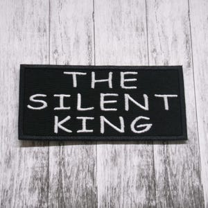 Op de afbeelding: Zwarte geborduurde patch met witte tekst die "THE SILENT KING" luidt.