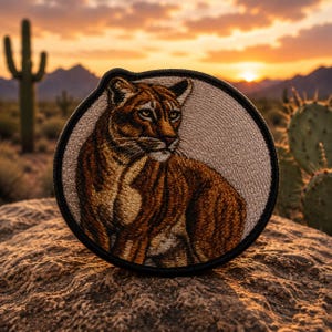 May include: Embroidered patch featuring a detailed cougar design in shades of brown and tan, set against a neutral background. The patch has a black border and is likely for application on clothing or accessories. The image is set against a desert sunset.