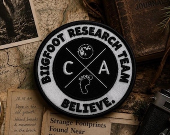 May include: A circular embroidered patch with a black center and a white border. The text "BIGFOOT RESEARCH TEAM" and "BELIEVE." is stitched in black. The center features a white cross design with the letters "C" and "A", a moon, and a footprint.