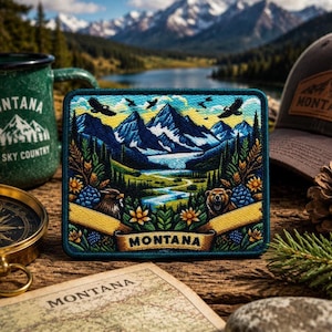 May include: A square patch with a Montana landscape design featuring mountains, a river, and wildlife. The patch has a teal border and a yellow banner with the word "MONTANA". A green mug and a hat with a Montana patch are also visible.