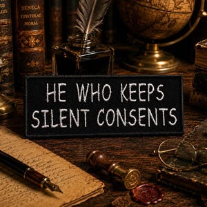 Op de afbeelding: Een rechthoekige zwarte geborduurde patch met de witte tekst HE WHO KEEPS SILENT CONSENTS. De patch heeft een strak gestikte rand en is ontworpen voor bevestiging op textiel, met duidelijke, leesbare letters in een vetgedrukt lettertype.