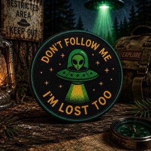 Könnte beinhalten: Ein schwarzer, gestickter Aufnäher mit dem Text "DON'T FOLLOW ME I'M LOST TOO" und einem Alien-Design in einem UFO. Der Aufnäher ist rund mit einem grünen Rand und gelbem Text. Der Hintergrund umfasst einen Kompass, einen Rucksack und ein Schild mit der Aufschrift "RESTRICTED AREA KEEP OUT".