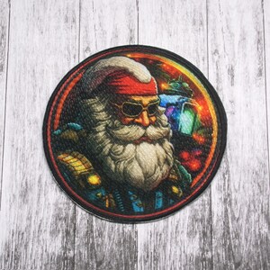 Puede incluir: Un parche redondo bordado con un Santa Claus de dibujos animados que lleva gafas de sol y un pañuelo rojo. El fondo es un diseño abstracto de colores.