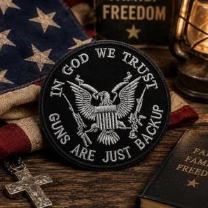 Könnte beinhalten: Ein runder, schwarzer, bestickter Aufnäher mit einem silbernen Adleremblem in der Mitte. Der Text IN GOD WE TRUST ist oben und GUNS ARE JUST BACKUP unten in weißen Großbuchstaben bogenförmig angeordnet.