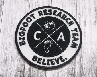 May include: Black and white embroidered patch with the text "Bigfoot Research Team Believe" around a circle containing a footprint, a cross, and the letters "C" and "A".