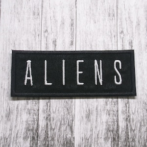 May include: Black fabric patch with white embroidered text that reads "ALIENS".