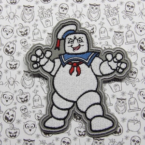 May include: A white and gray embroidered patch of a ghost with a blue sailor collar and red bow tie. The ghost has a mischievous expression and is wearing a sailor hat. The patch is on a white background with a Halloween pattern.