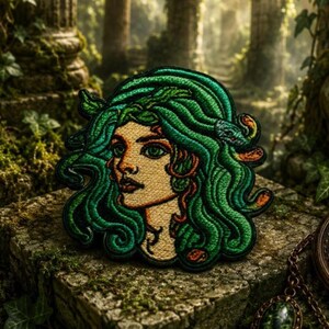 May include: An embroidered patch featuring a stylized portrait of a figure with flowing green hair composed of intertwined snakes. The design uses shades of emerald and forest green thread against a light tan skin tone, with dark outlines.