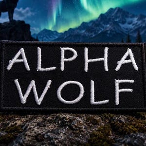 Pode incluir: Um patch retangular preto com as palavras "ALPHA WOLF" bordadas em branco. O patch é colocado em um fundo de uma silhueta de lobo, montanhas e aurora boreal. O patch é provavelmente para uma jaqueta ou bolsa.