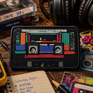 May include: A black patch embroidered with a colorful cassette tape design. The patch features red, blue, orange, and green accents. Other items in the image include cassette tapes, headphones, and a notepad with the word "mixtape".