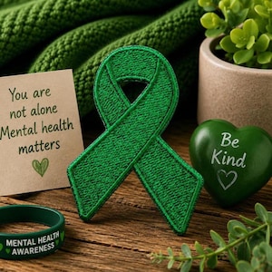 Op de afbeelding: Een groen lint, een hartvormig object met de tekst "Be Kind" en een armband met de tekst "Mental Health Awareness" liggen op een houten oppervlak. Een kaart met de tekst "You are not alone Mental health matters" is ook aanwezig. Een kleine plant staat op de achtergrond.