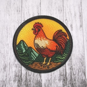 May include: A round embroidered patch with a rooster standing on a green hill. The rooster is red, orange, and white with a yellow background. The patch is surrounded by a black border.