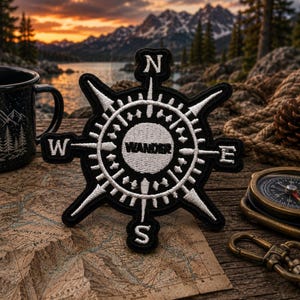May include: A black and white embroidered patch featuring a compass rose design with the word WANDER in the centre. The patch includes the cardinal directions N, S, E, and W in white stitching against a black background with detailed needlework.